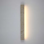 A vertical Clérachic travertine wall light with a warm glow down one side, against a soft grey-blue wall.