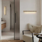 A Clérachic travertine wall light hangs on a white wall beside a tall reeded door, above a boucle armchair in the corner.