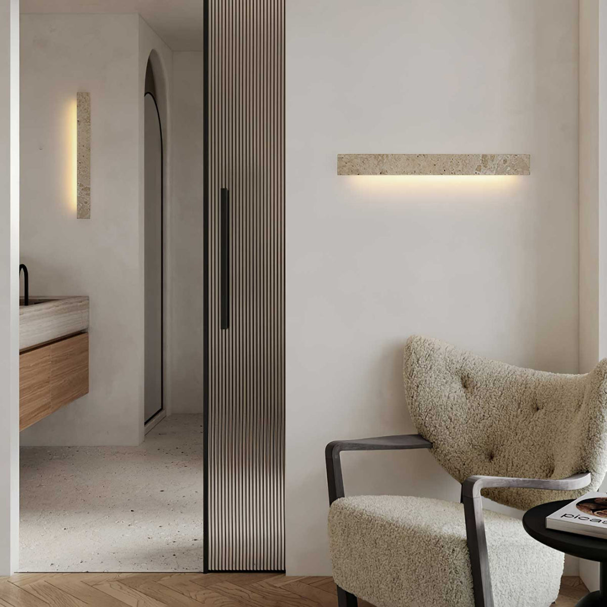 A Clérachic travertine wall light hangs on a white wall beside a tall reeded door, above a boucle armchair in the corner.