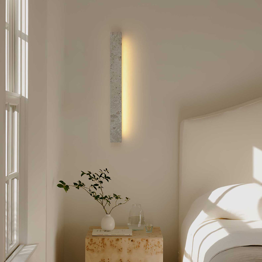 A vertical Clérachic travertine wall light glows warm beside a window, above a stone side table and a bed with white linen.