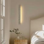 A vertical Clérachic travertine wall light glows warm beside a window, above a stone side table and a bed with white linen.