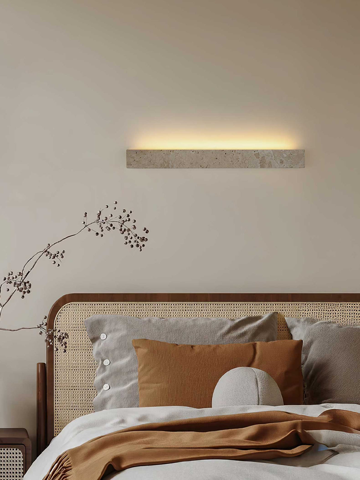 A Clérachic travertine wall light glows above a rattan bed with grey and rust cushions and a dried branch.