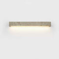 A horizontal Clérachic wall light in speckled travertine with a warm glow beneath, on a pale backdrop.