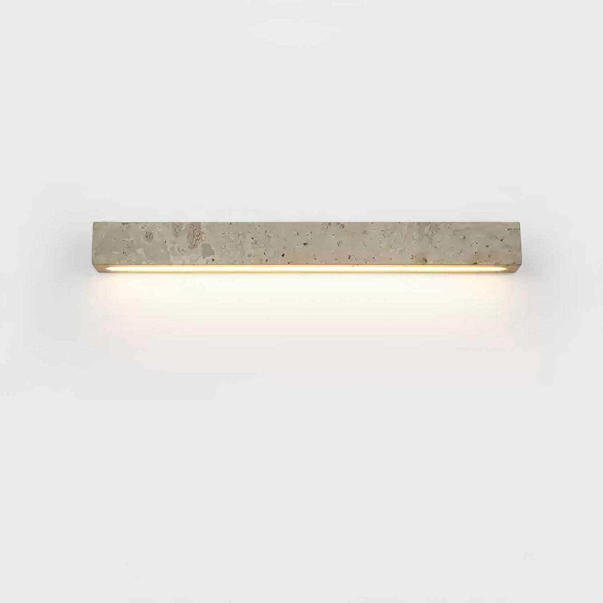 A horizontal Clérachic wall light in speckled travertine with a warm glow beneath, on a pale backdrop.