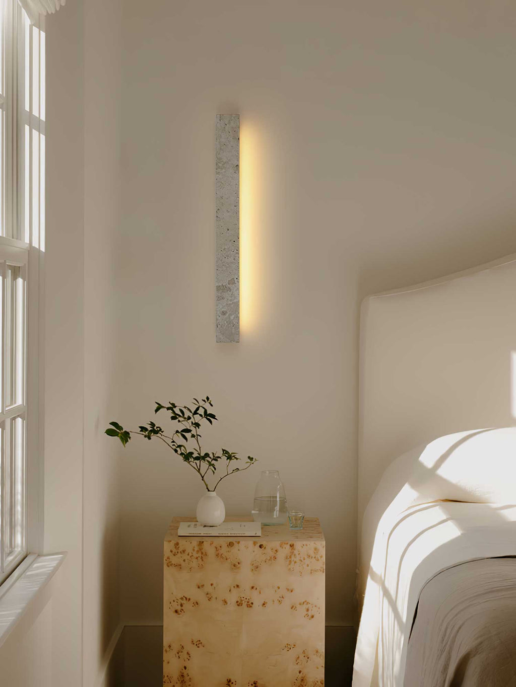 A tall Clérachic travertine wall light glows beside a window, above a burr wood bedside cube with a small plant.