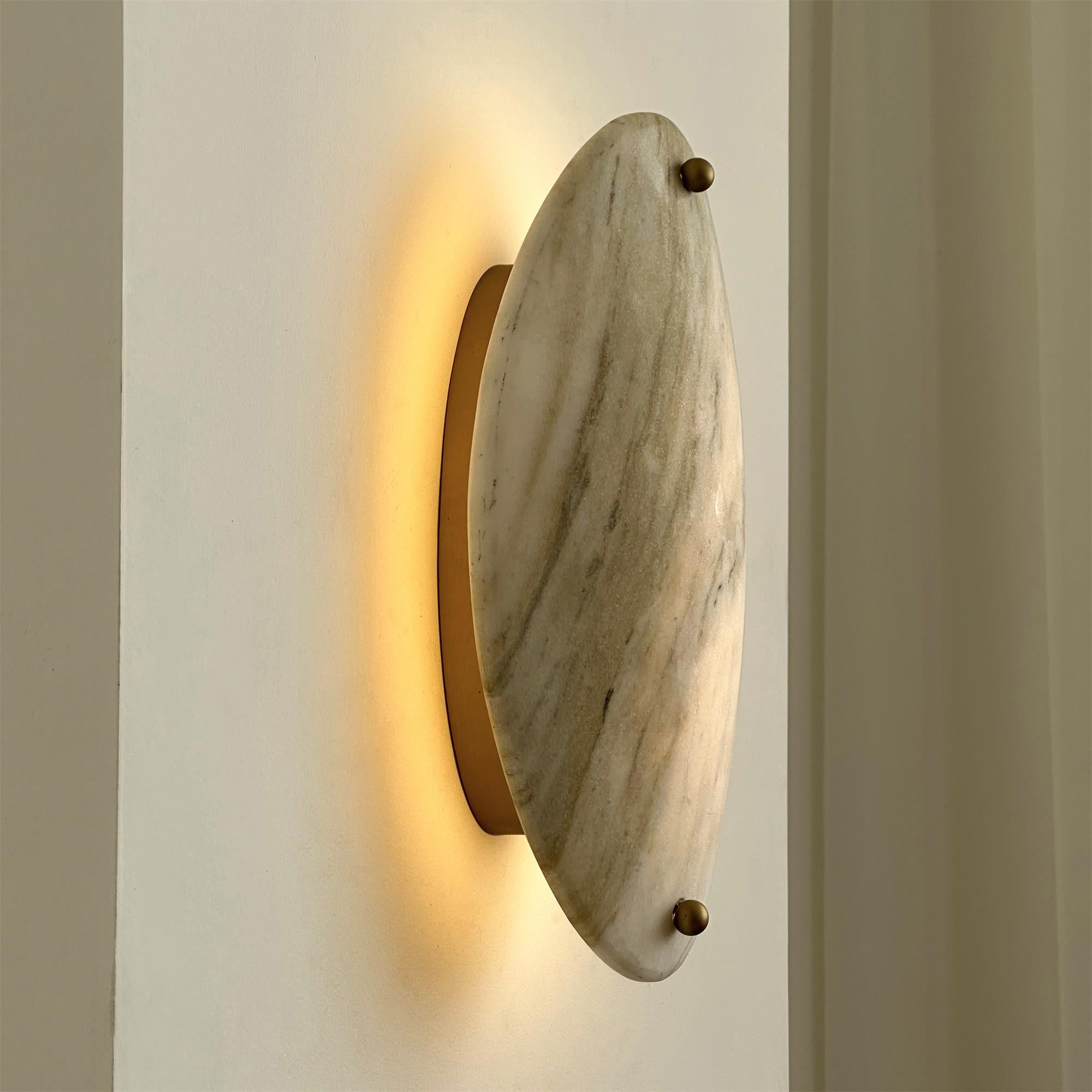The Clérachic sconce lit at an angle, warm light spreading up and down the wall past the brass mounting disc.