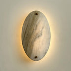 Clérachic wall lamp lit head-on, a warm ring of light behind the oval bringing out the veining in the stone.