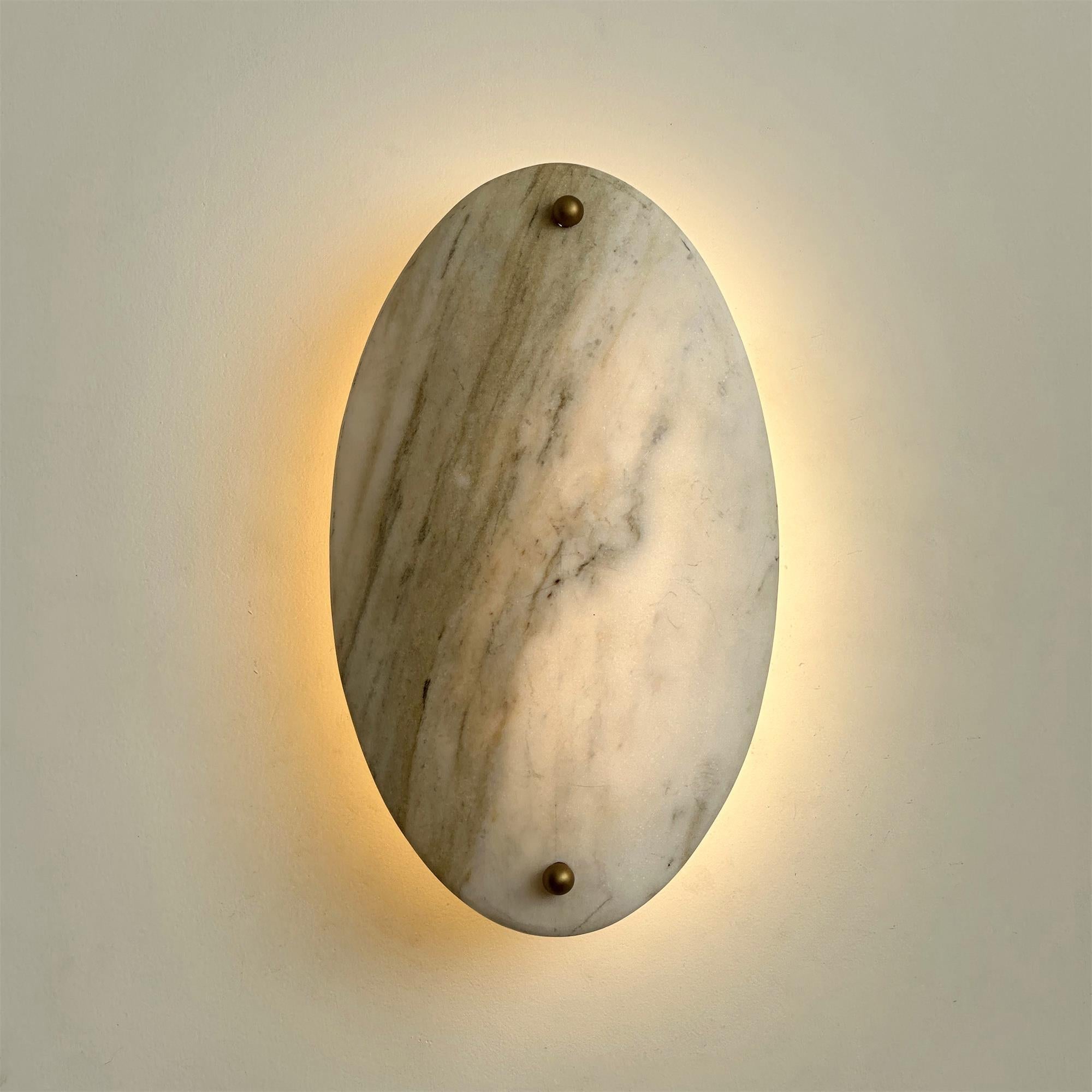 Clérachic wall lamp lit head-on, a warm ring of light behind the oval bringing out the veining in the stone.