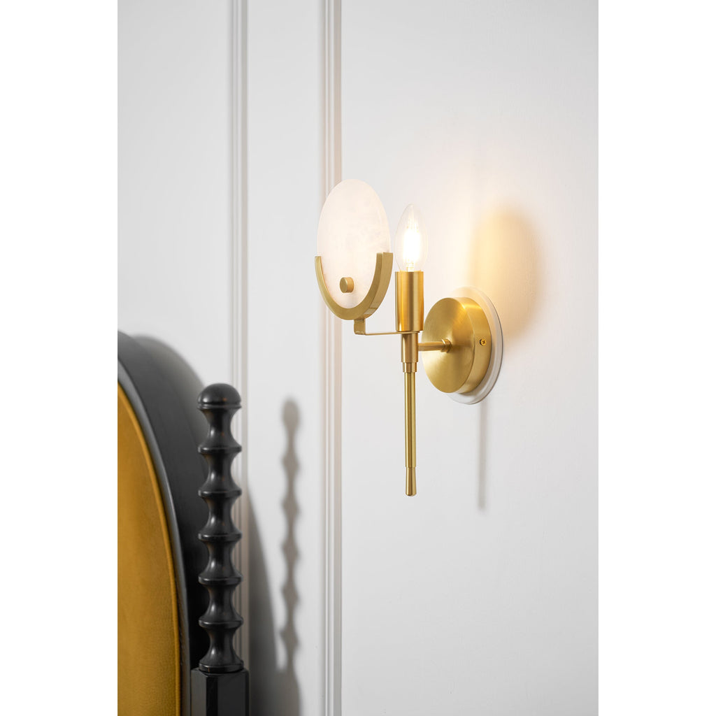 Clérachic brass wall light in close-up, its alabaster disc and lit candle bulb set against white panelled bedroom wall.
