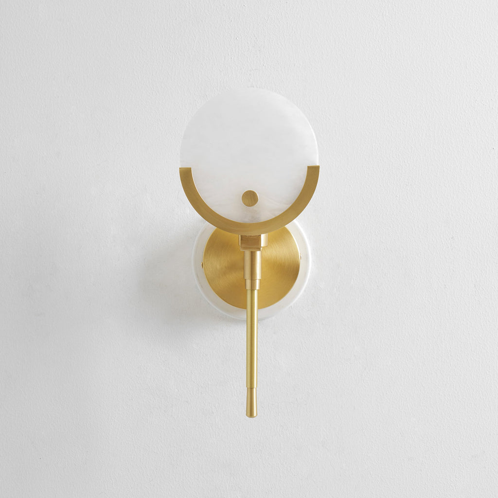 The Clérachic wall light on plain white plaster, its round alabaster shade cradled in a brushed brass crescent.