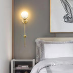 The Clérachic sconce lit in a grey bedroom corner, brass stem running down beside a gold-framed print.