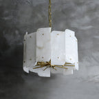Unlit Clérachic Zoe alabaster chandelier on its brass chain against a raw concrete wall, every stone panel veined differently.