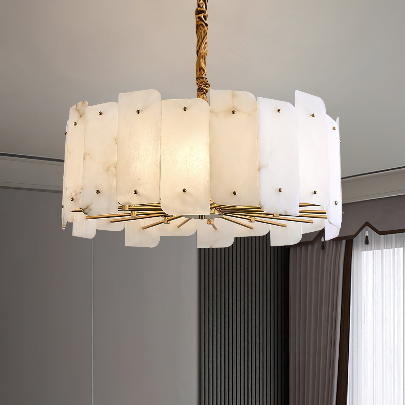 The Clérachic Zoe alabaster chandelier lit in a grey panelled room, hanging low from its brass stem beside floor-length curtains.