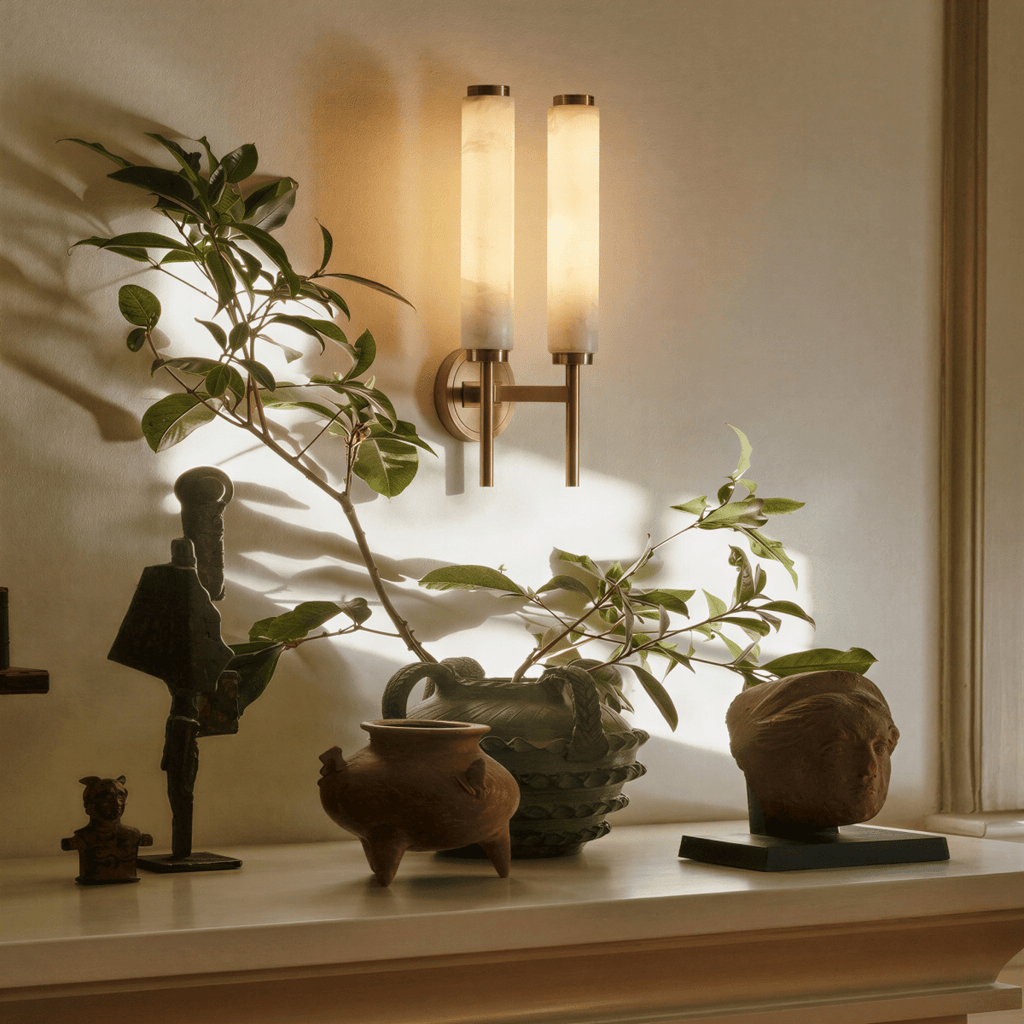 Clérachic Colen two-head wall lamp in gold lit above a shelf arranged with a trailing plant, terracotta vessels and a weathered stone head.