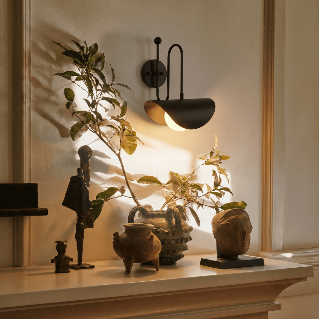 Clérachic matte black curved-arm wall sconce lit above a mantel with carved wooden figures and pottery.