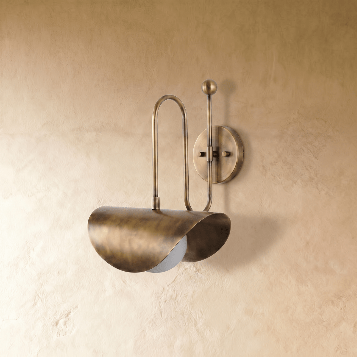 Clérachic curved-arm wall sconce in vintage brass, shown from a slightly closer studio angle on a plaster wall.