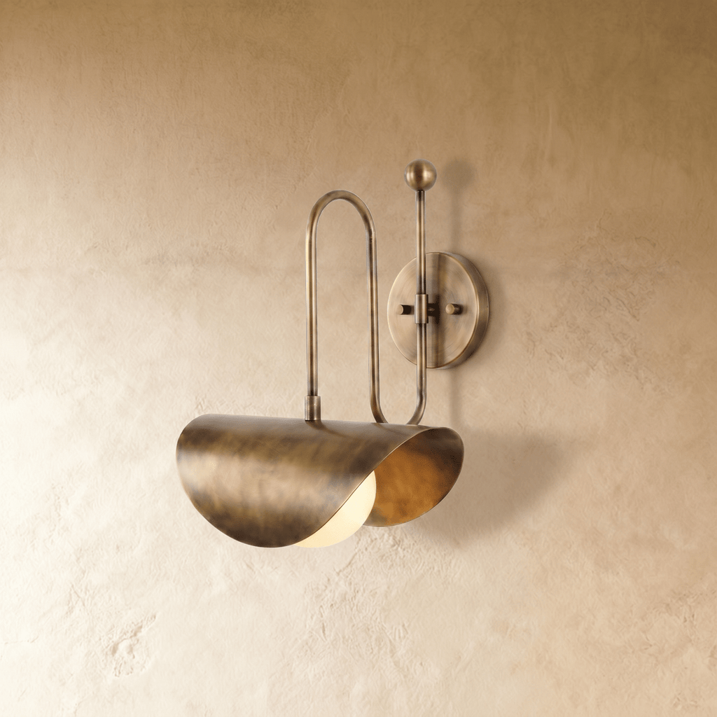 Clérachic curved-arm wall sconce in vintage brass with a folded scallop shade, mounted on a plaster wall in studio light.