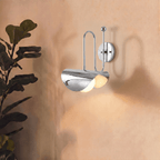 Clérachic polished chrome curved-arm wall sconce lit on a plain wall beside a leafy fiddle-leaf fig plant.
