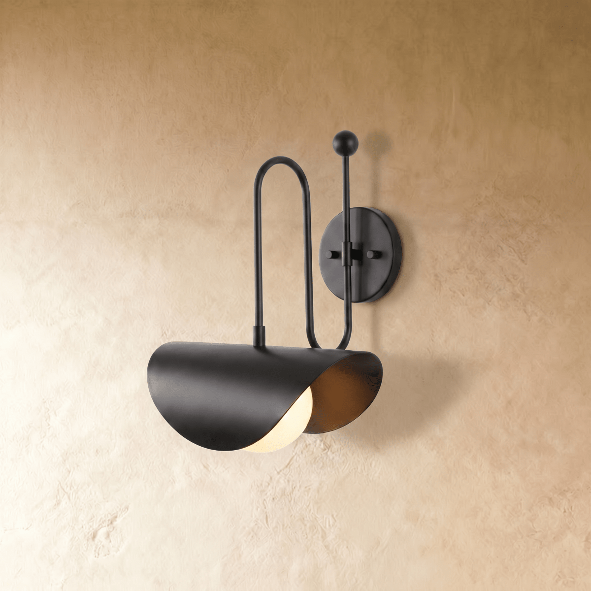 Clérachic curved-arm wall sconce in matte black with a folded scallop shade, mounted on a plaster wall in studio light.
