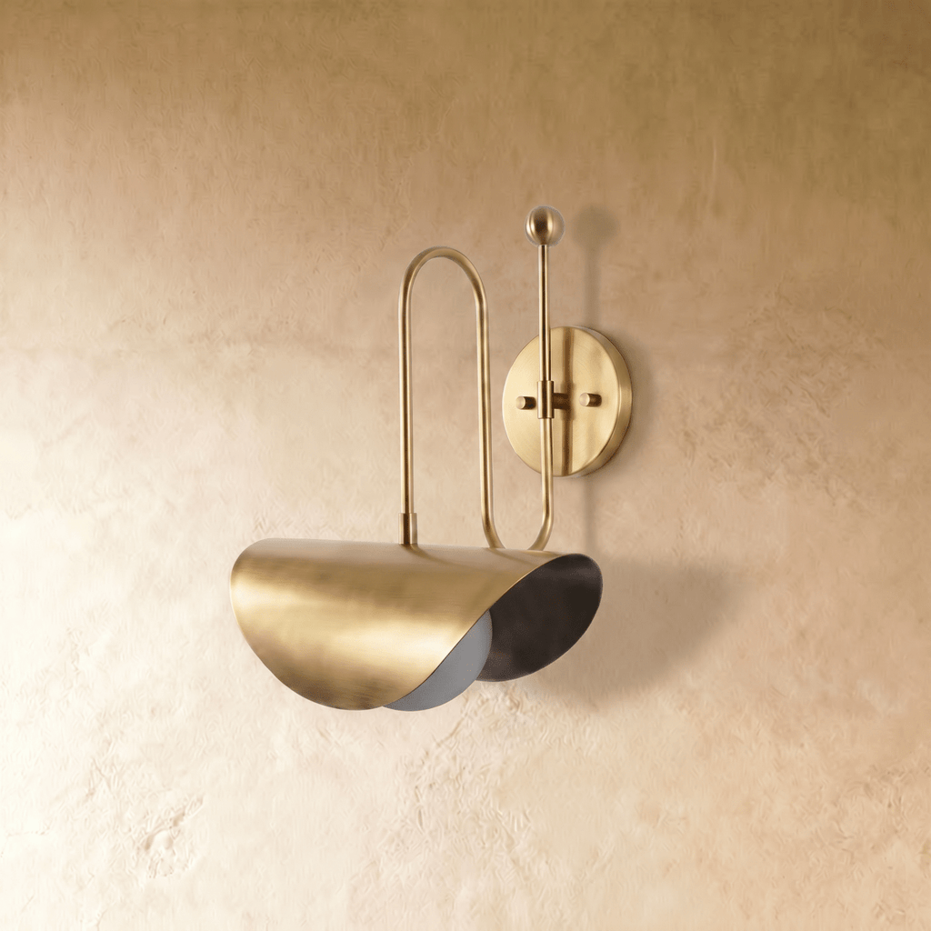 Clérachic curved-arm wall sconce in polished brass with a folded scallop shade, mounted on a plaster wall in studio light.