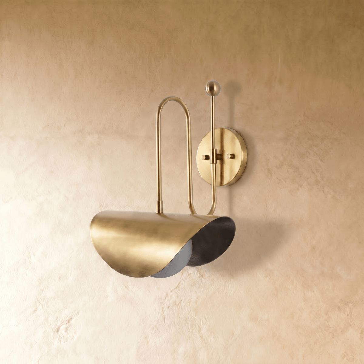 Clérachic curved-arm wall sconce in polished brass with a folded scallop shade, mounted on a plaster wall in studio light.