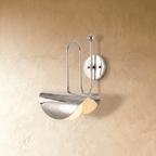 Clérachic curved-arm wall sconce in polished chrome with a folded scallop shade, mounted on a plaster wall in studio light.