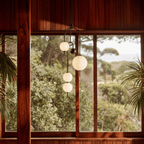 Clérachic pendant light in its four-globe build, hanging in front of a tall timber-framed window that looks out over dense trees.