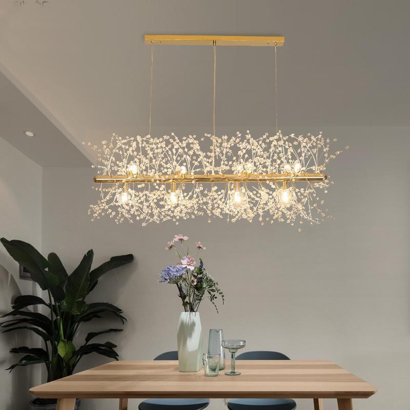 A rectangular gold-bar pendant with crystal-tipped branches hangs above a wood table set with dried flowers, a different Clérachic fixture from this chandelier.