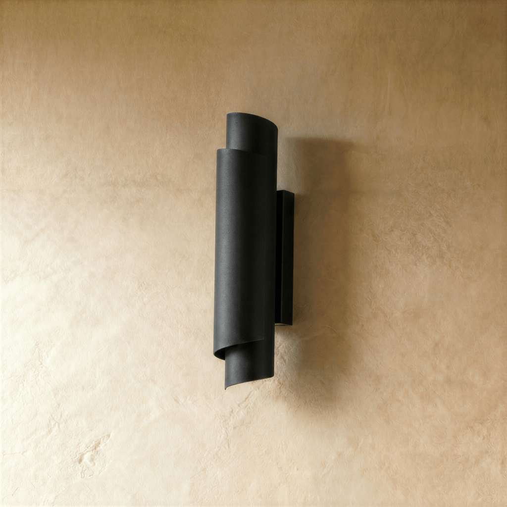 Clérachic wall sconce in matte black, the curled metal form mounted on a pale wall with light spilling up and down.