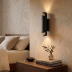 Clérachic matte black wall sconce lit above a wooden nightstand stacked with books and a small glass vase in a bedroom.