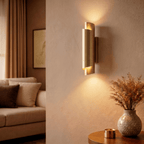 Clérachic brass wall sconce lit above a round side table with a patterned vase and dried grasses in a living room.