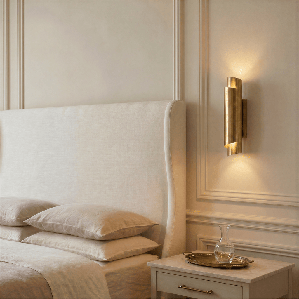 Clérachic brass wall sconce glowing beside an upholstered headboard, above a nightstand with a glass carafe on a tray.