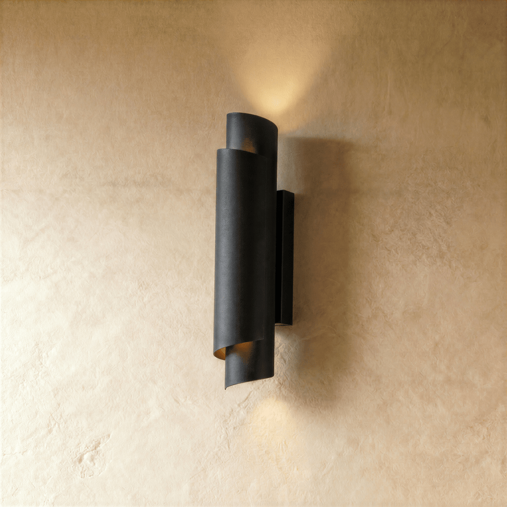 Clérachic wall sconce in matte black, its rolled cylinder shade casting a warm glow up and down a plaster wall.