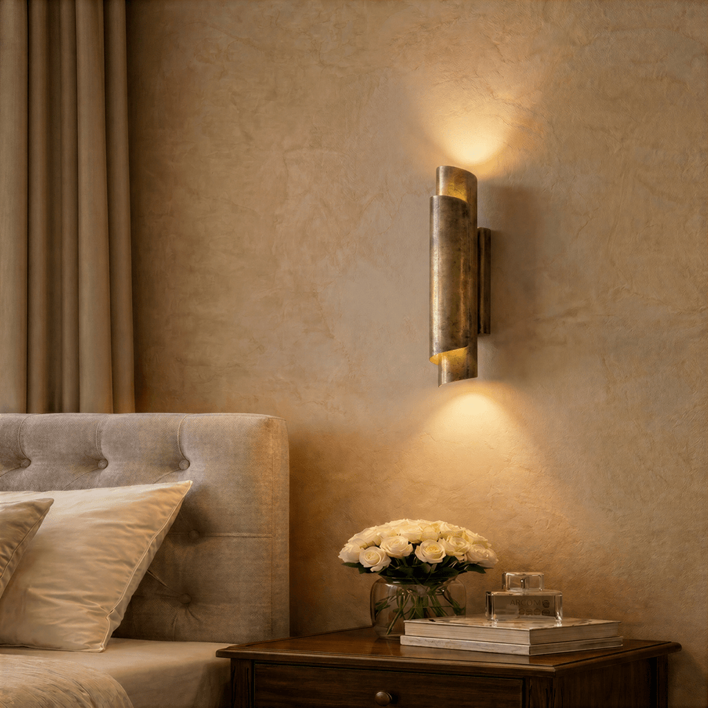 Clérachic vintage brass wall sconce lit above a bedside table with white roses and books, beside an upholstered headboard.