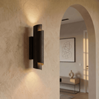 Clérachic matte black wall sconce glowing in a plaster hallway beside an archway leading to a sunlit living room.