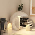 Lit on a white desk corner, the Clérachic resin crescent lamp sits beside a stack of books, a wire magazine rack and a pale mug.
