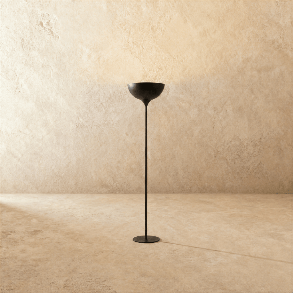 Clérachic floor lamp in matte black, its shallow uplight bowl set on a slender stem against a plaster wall.