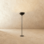 Clérachic floor lamp in matte black, its shallow uplight bowl set on a slender stem against a plaster wall.