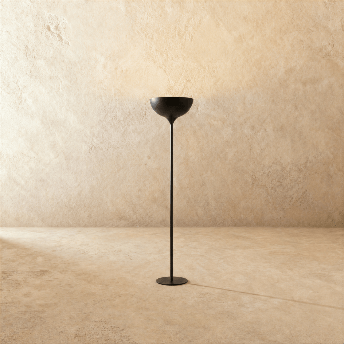 Clérachic floor lamp in matte black, its shallow uplight bowl set on a slender stem against a plaster wall.