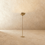 Clérachic floor lamp in a brighter brass finish, its uplight bowl set on a slender stem against a plaster wall.