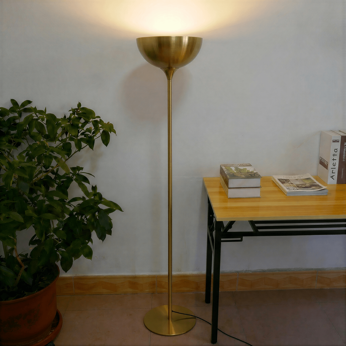 Clérachic brass floor lamp glowing beside a potted plant and a small table stacked with books and magazines.
