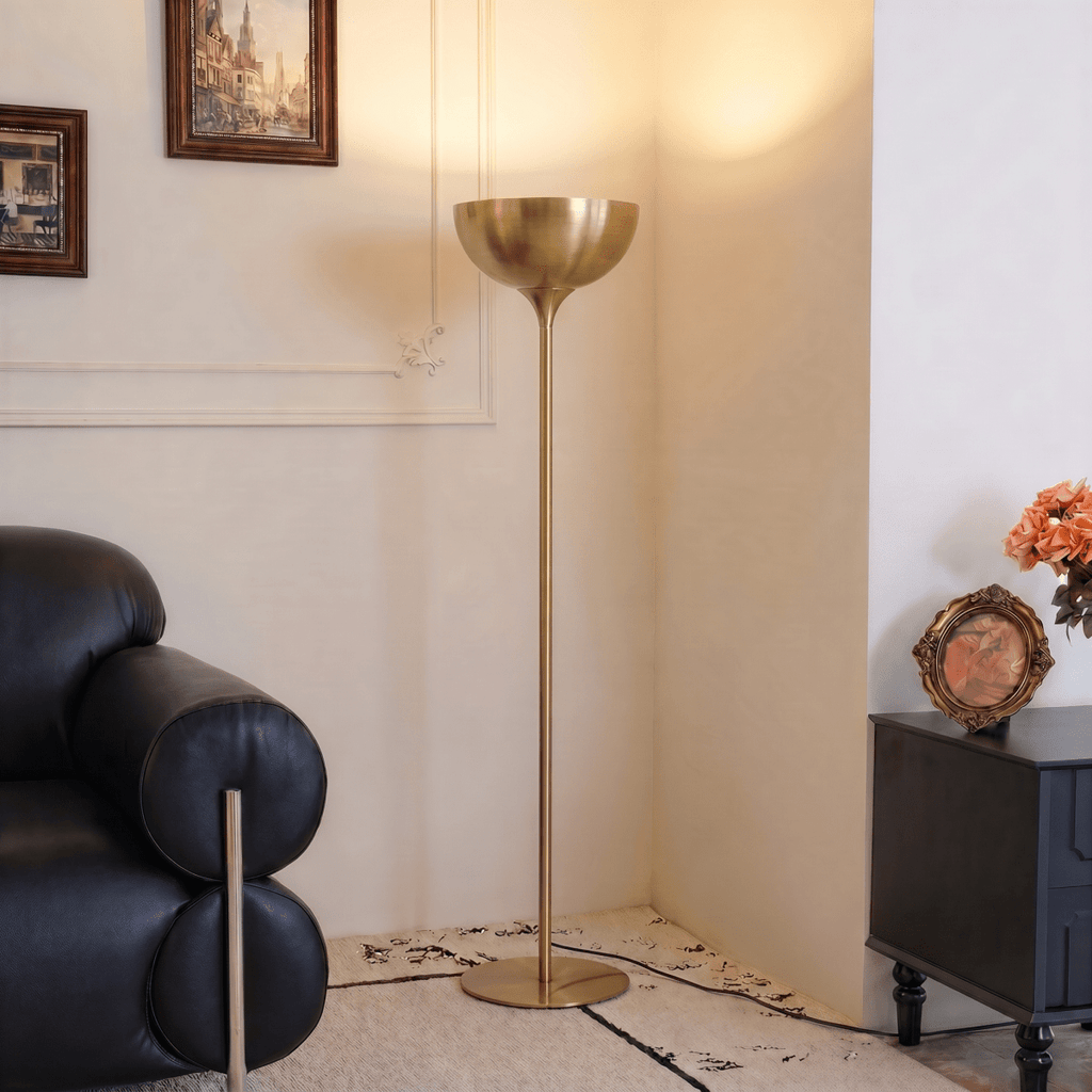Clérachic brass floor lamp glowing beside a black leather sofa and a gilt-framed painting near a patterned rug.