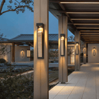 Clérachic wall lamp repeated down a row of columns under a slatted pergola walkway, each one lit at dusk.
