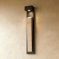 Clérachic travertine wall lamp lit on a warm-toned textured wall, the light in its canopy washing down the stone's face.