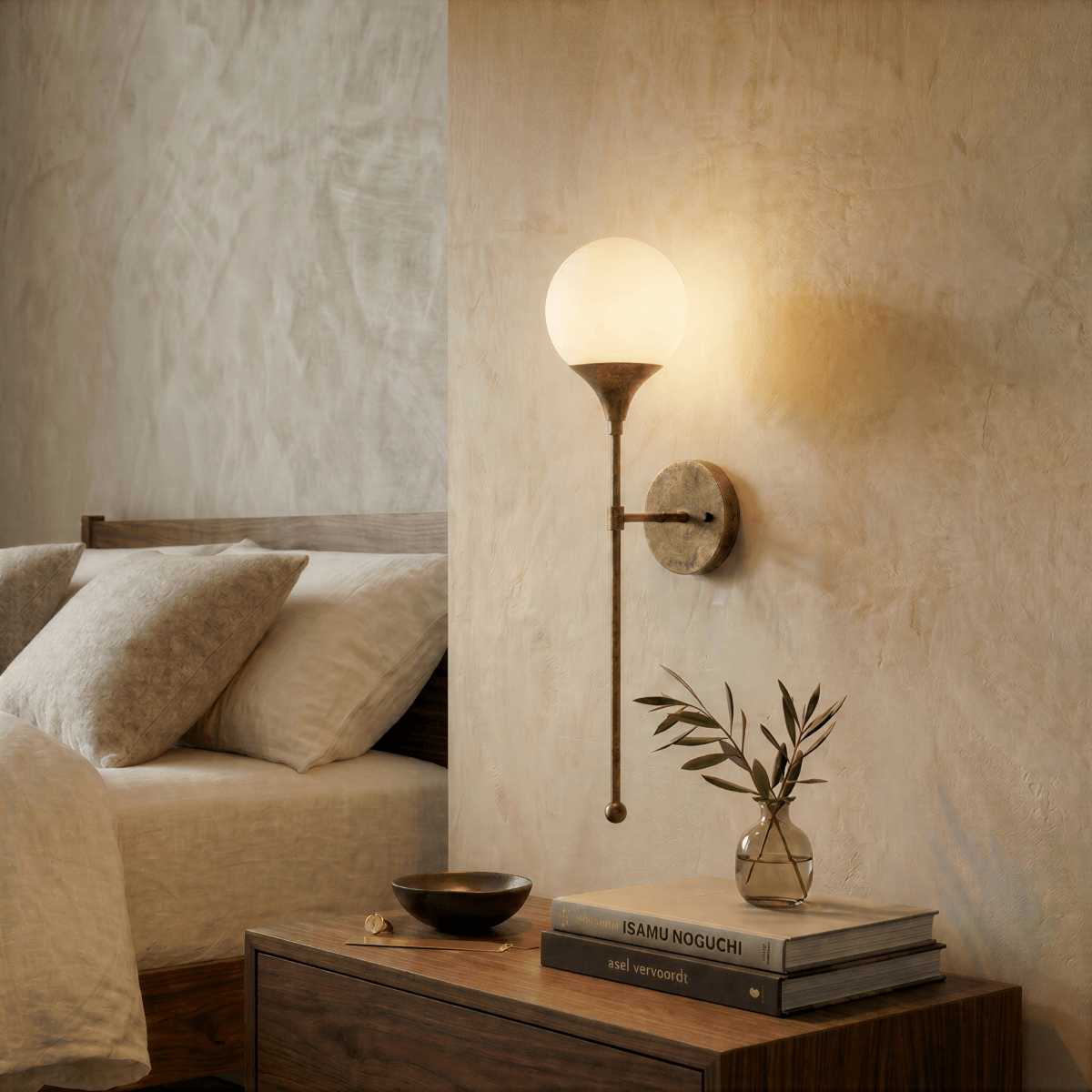 Clérachic wall lamp in brass glowing over a nightstand stacked with books and a sprig of olive.