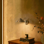 Clérachic chrome cone-shade wall sconce lit above a bedside table with stacked books and spring blossoms.