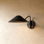 Clérachic cone-shade wall sconce in matte black with a curved arm, mounted on a plaster wall in studio light.