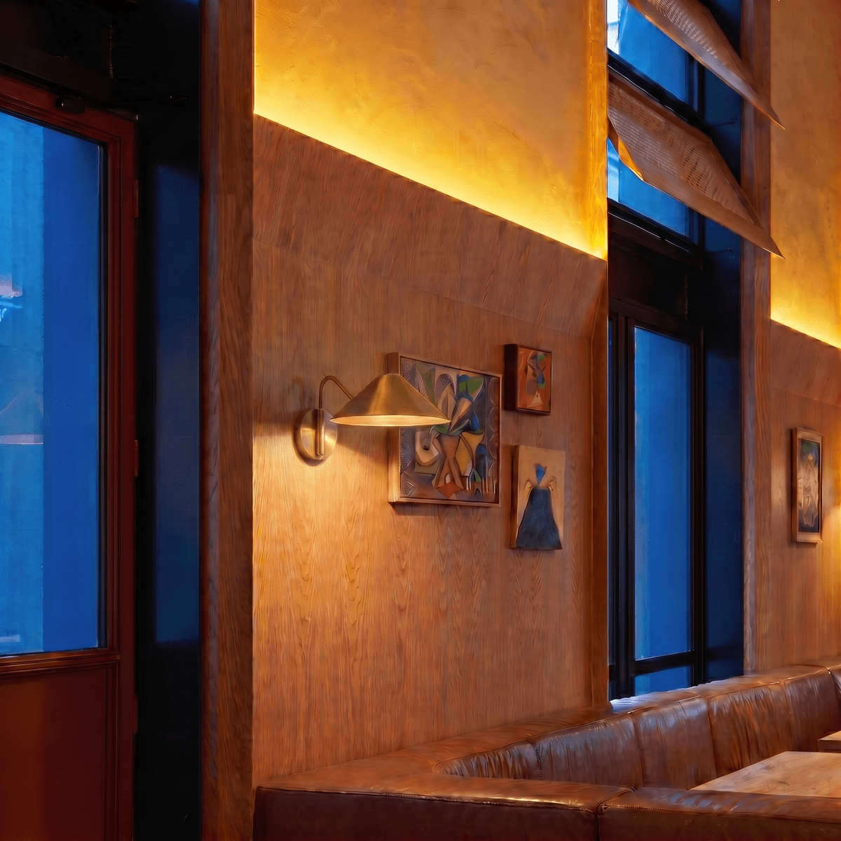 Clérachic brass cone-shade wall sconce lit on a wood-panelled wall beside small framed artworks in a lounge.