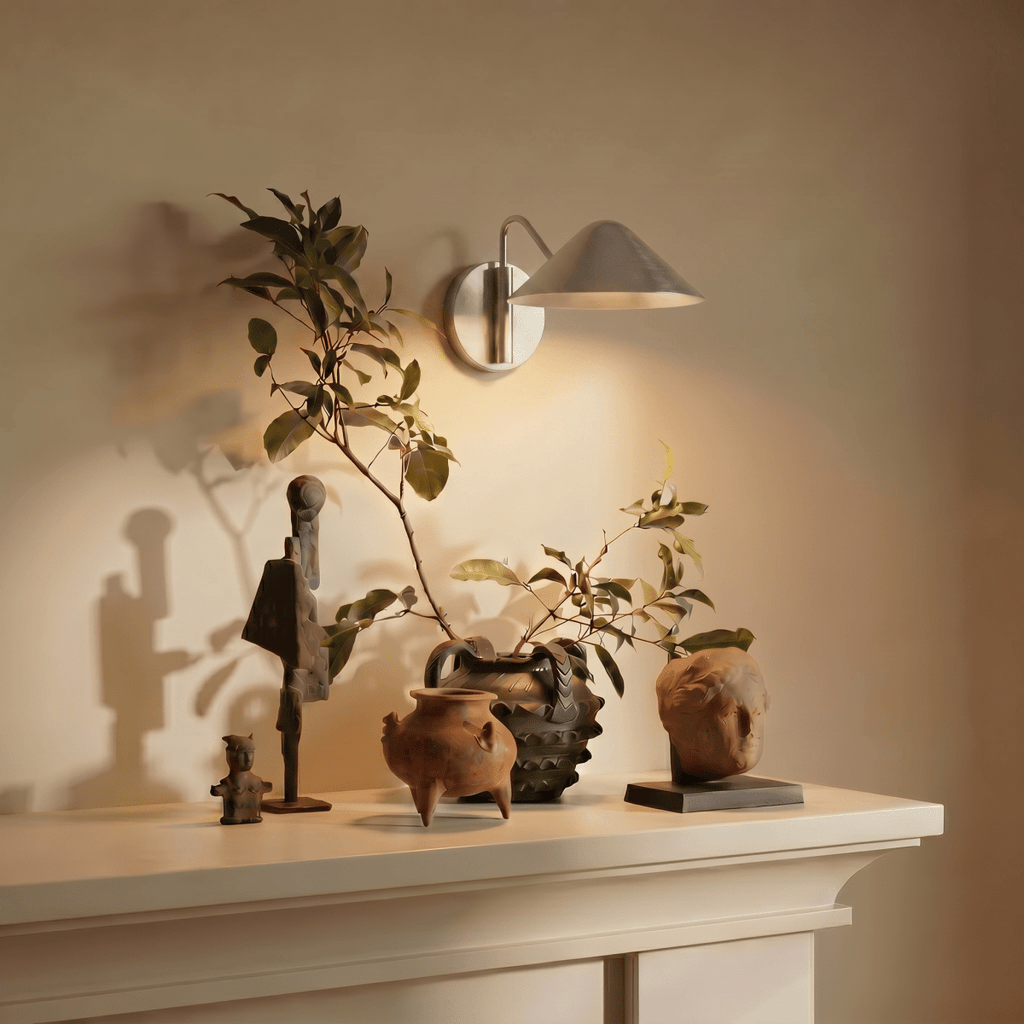 Clérachic chrome cone-shade wall sconce lit above a mantel displaying carved wooden figures and pottery.