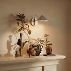 Clérachic chrome cone-shade wall sconce lit above a mantel displaying carved wooden figures and pottery.